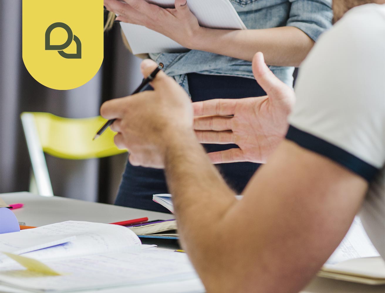 Ein Mann gestikuliert mit den Händen, während er einen Bleistift hält. Vor ihm ist ein Tisch mit einigen aufgeklappten Büchern. Über dem Bild befindet sich ein Kommunikationssymbol. Das Symbol zeigt zwei Sprechblasen.